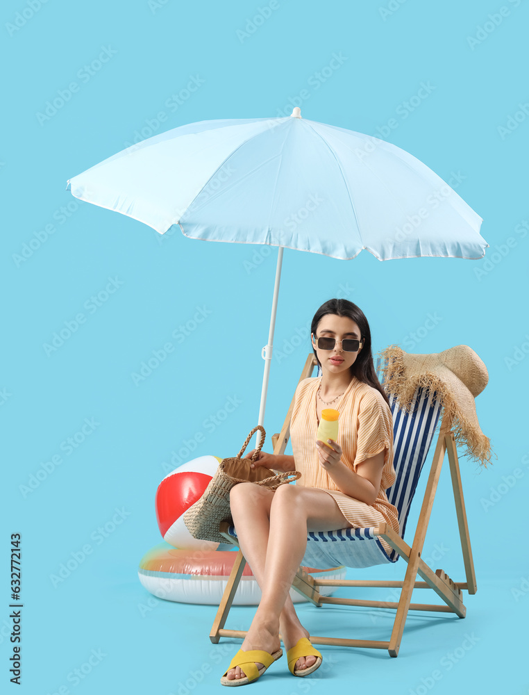 Young woman with sunscreen cream and beach bag in deck chair on blue background