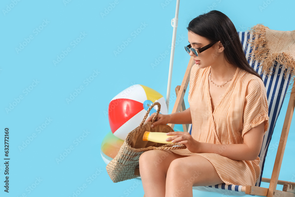 Young woman with sunscreen cream and beach bag in deck chair on blue background