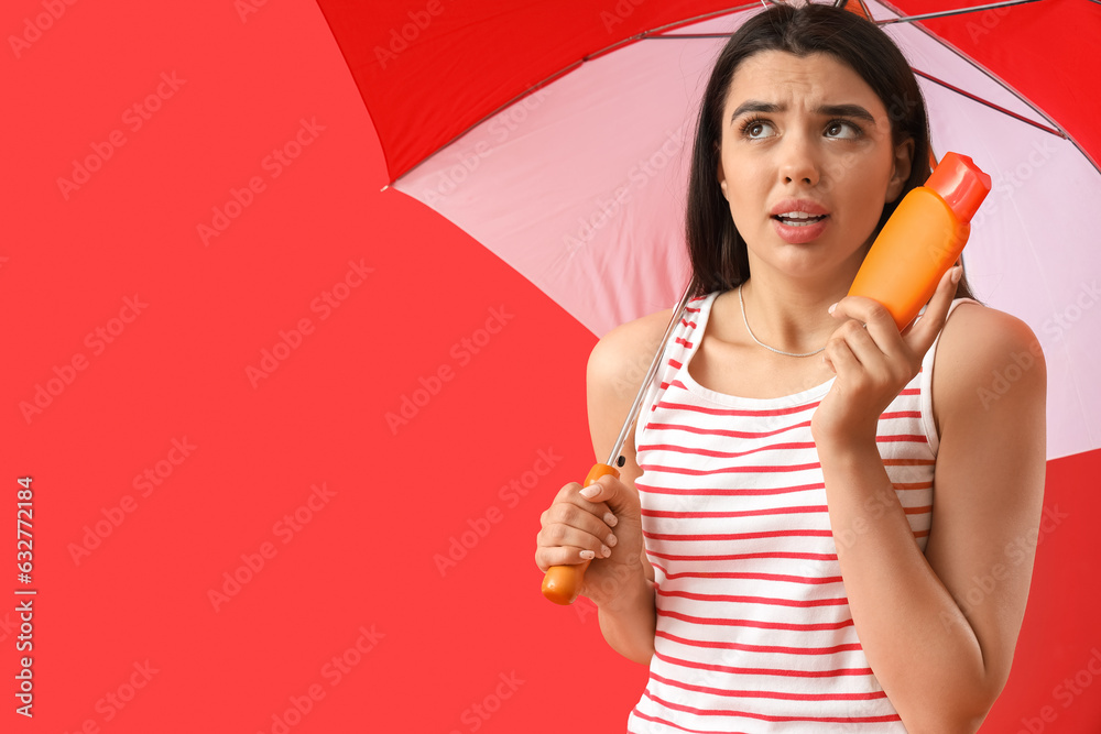 Worried young woman with sunscreen cream and beach umbrella on red background