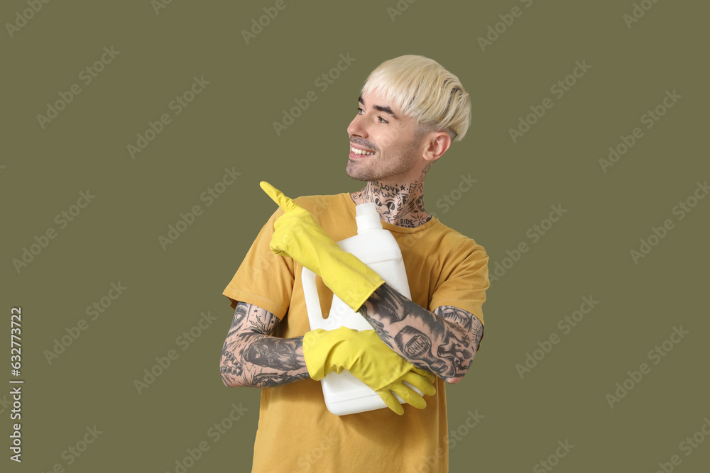 Young tattooed man with bottle of detergent pointing at something on green background