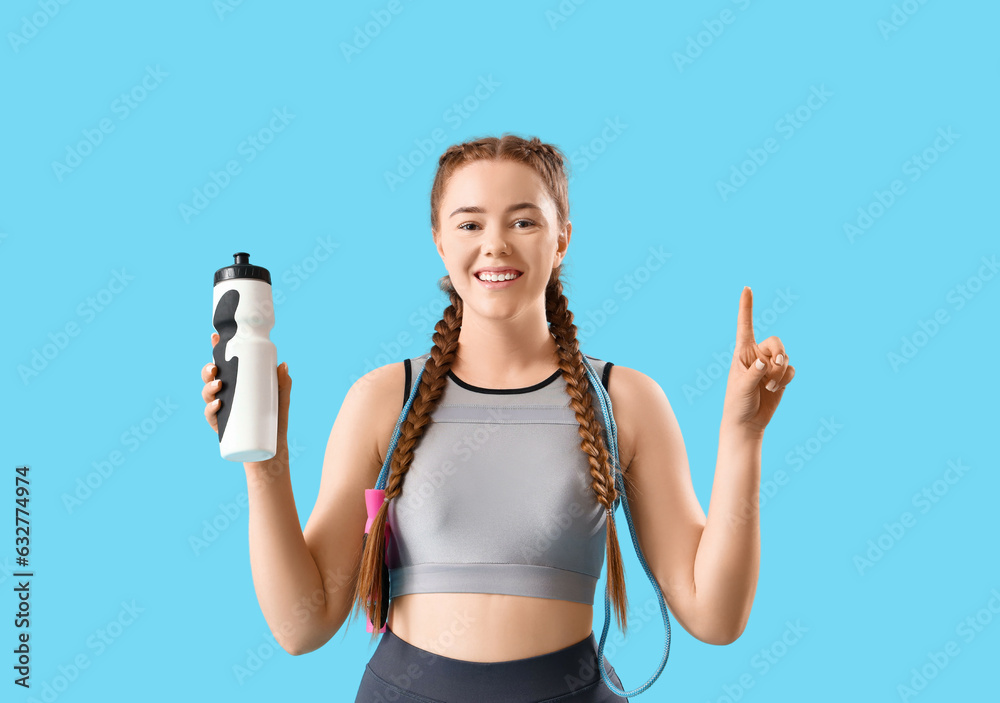 Sporty young woman with skipping rope and bottle pointing at something on blue background