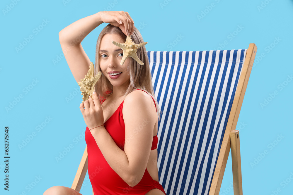 Beautiful young woman with starfishes sitting in deck chair on blue background