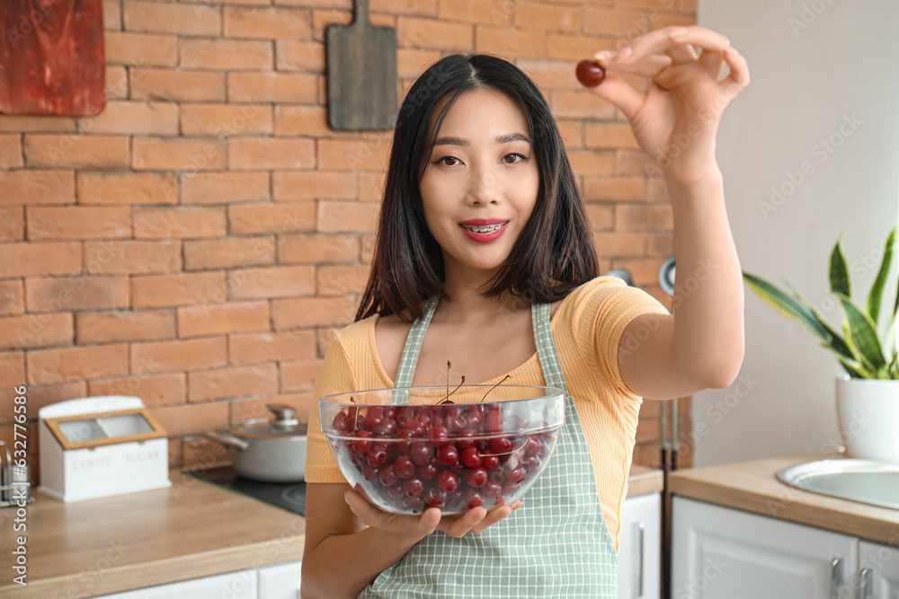 Happy beautiful young Asian woman in apron with bowl of ripe cherries in kitchen