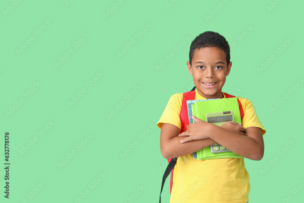 Little African-American boy with backpack and school supplies on green background