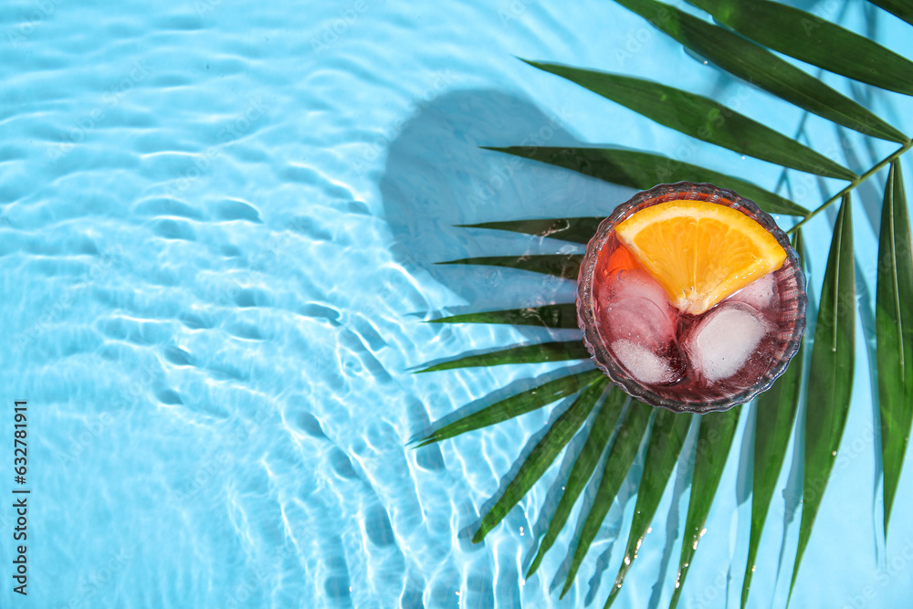 Glass of cold Negroni cocktail and palm leaf in water on color background