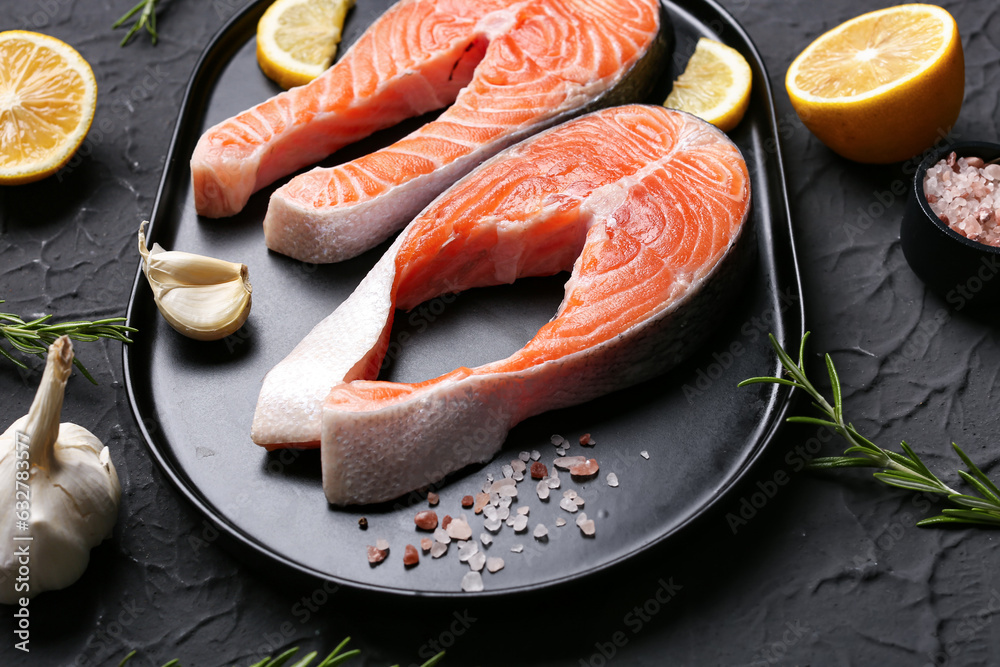 Plate with raw salmon steaks and different spices on black background