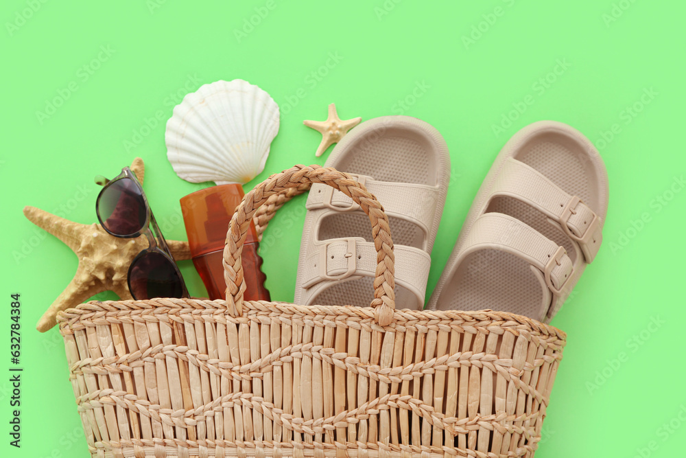 Pair of beige flip flops and straw bag with beach accessories on green background