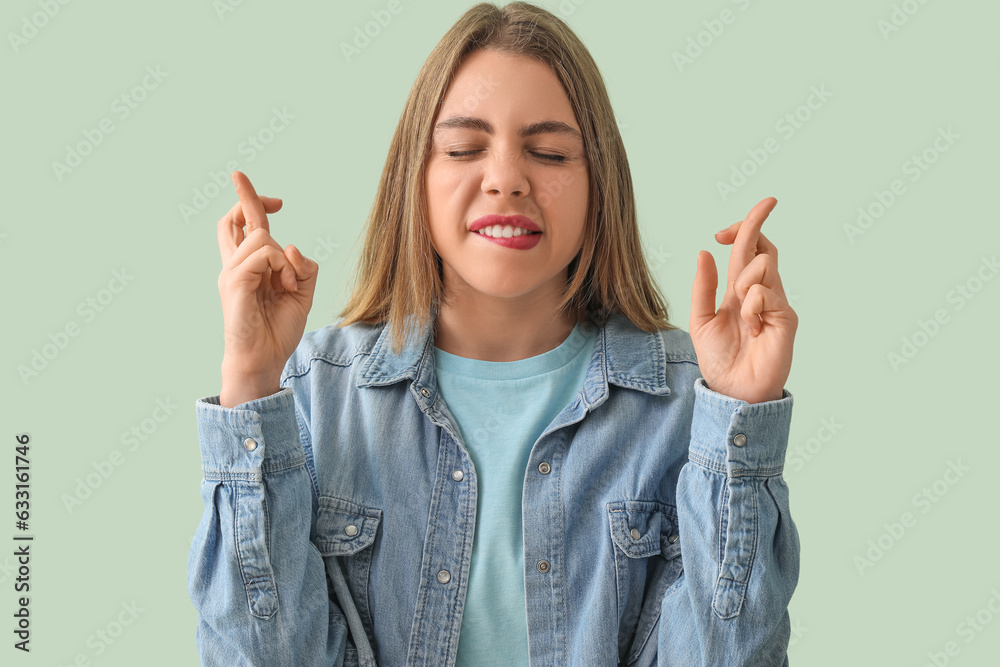 Young woman in denim shirt crossing fingers on green background, closeup