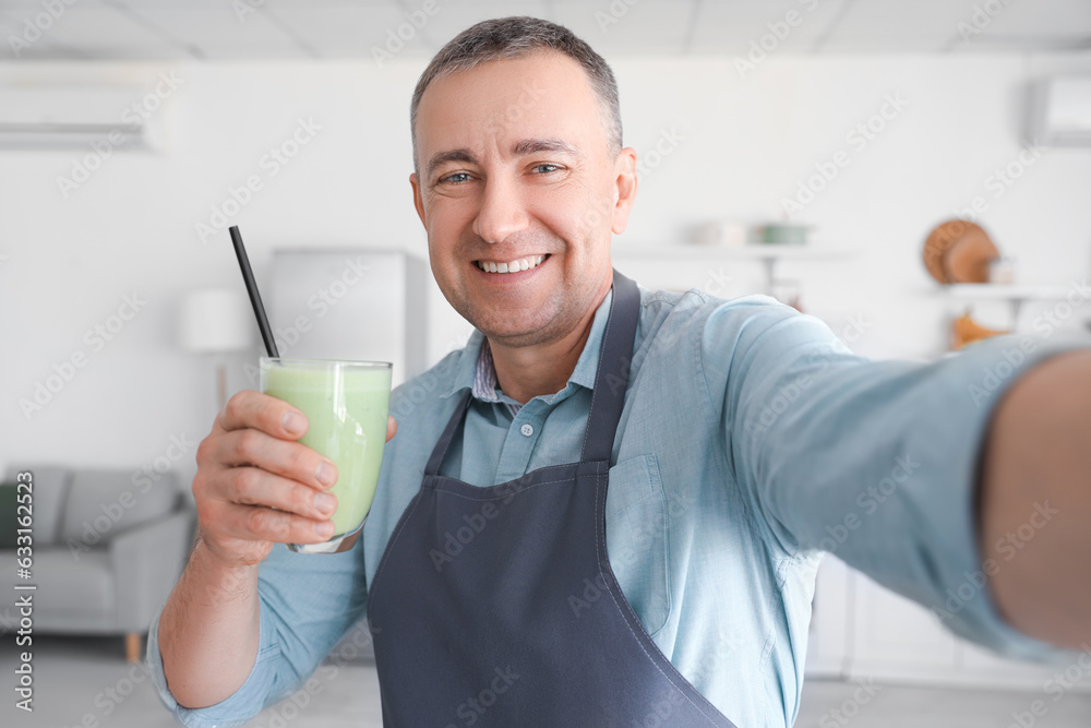 Mature man with glass of fresh vegetable smoothie taking selfie in kitchen