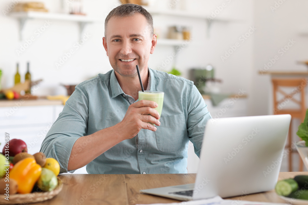 Mature man with glass of fresh vegetable smoothie using laptop in kitchen