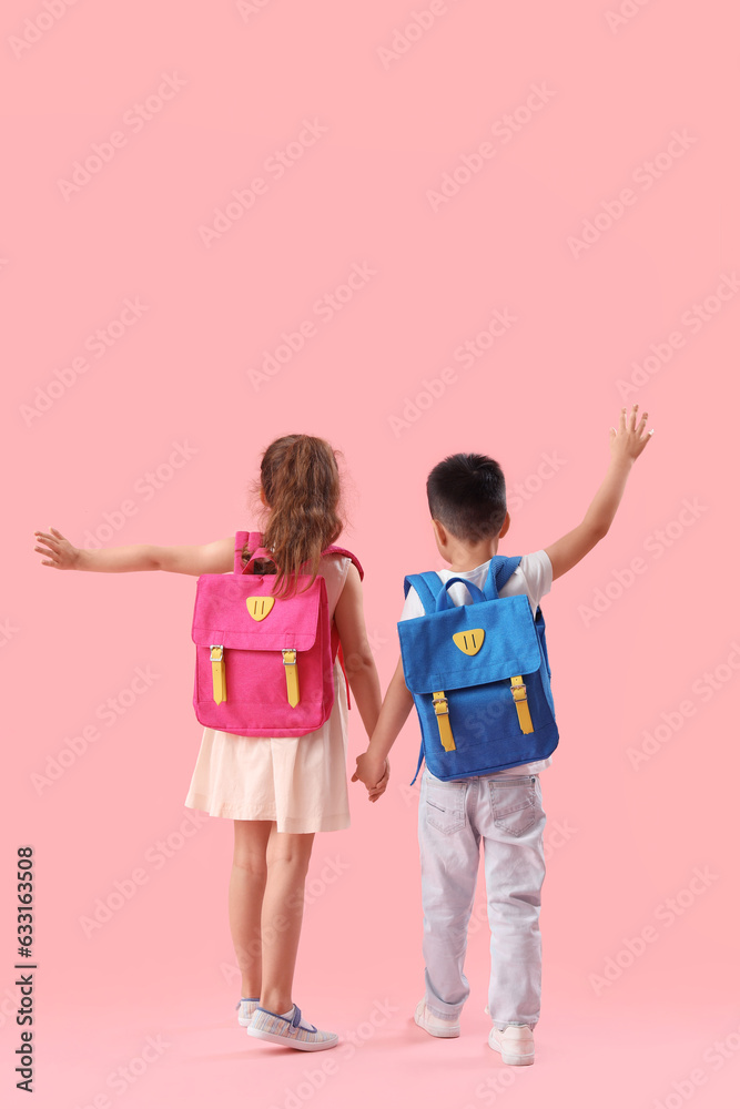 Cute little pupils with backpacks holding hands on pink background, back view