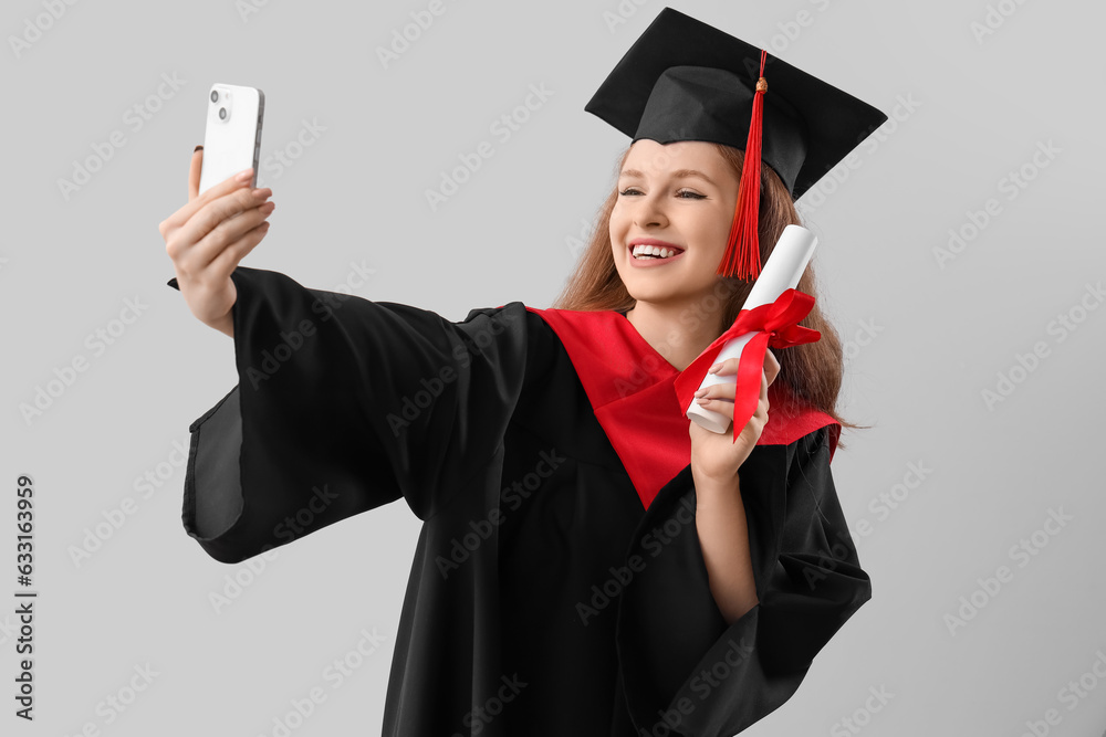 Female graduate student with diploma taking selfie on grey background