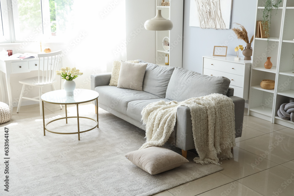 Cozy grey sofa with soft blanket and coffee table in interior of light living room