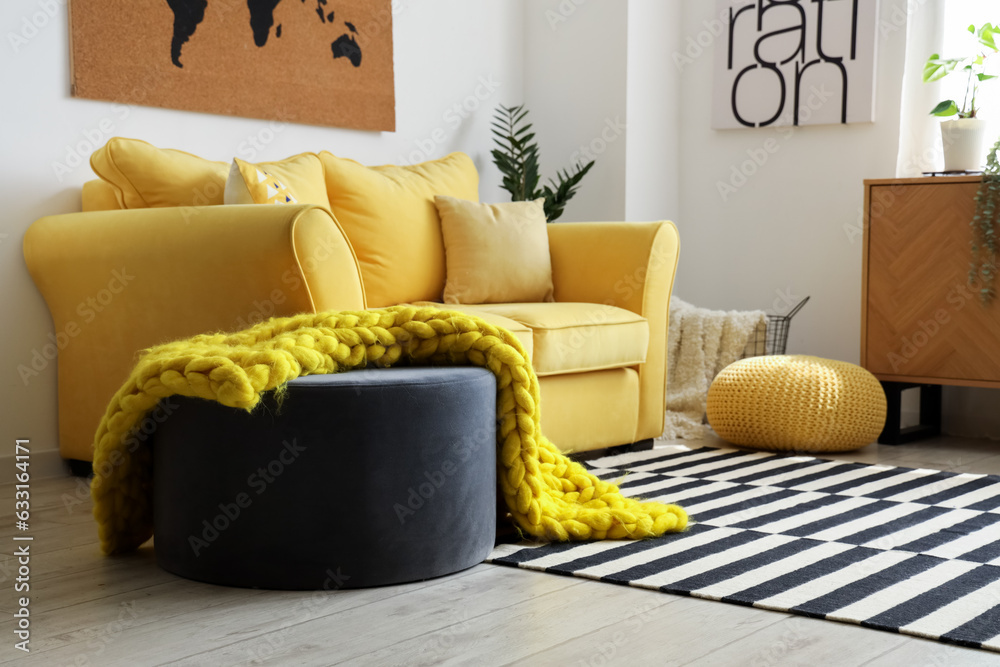 Interior of stylish living room with cozy yellow sofa and soft knitted blanket on pouf