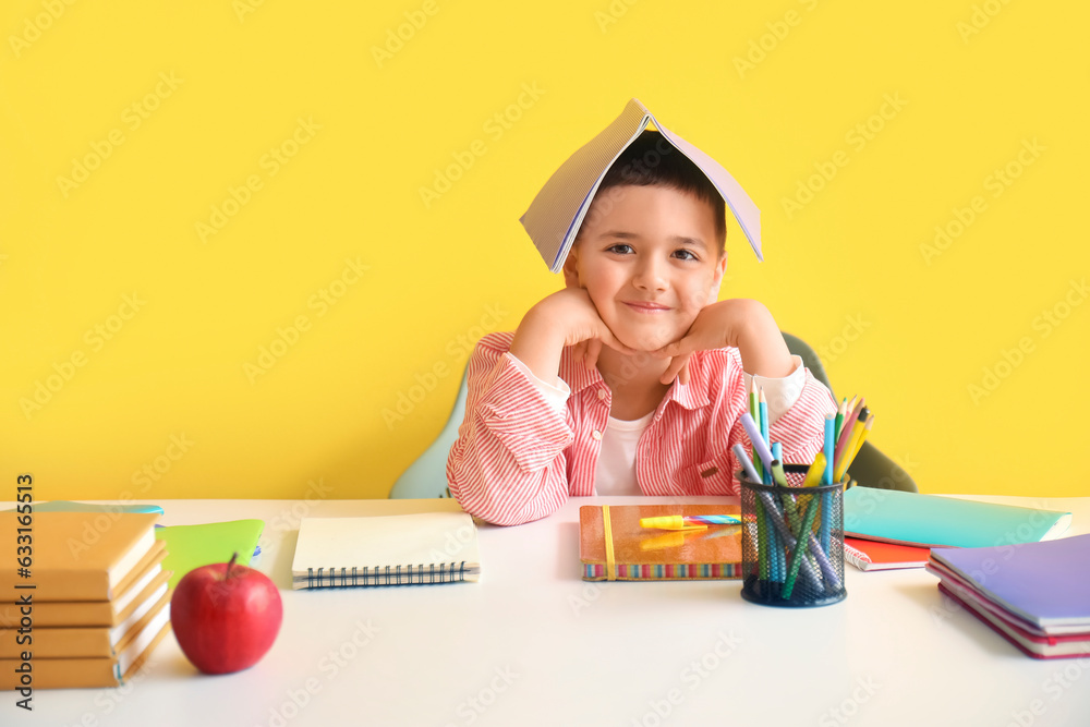 Cute little boy with copybook doing homework at table near yellow wall