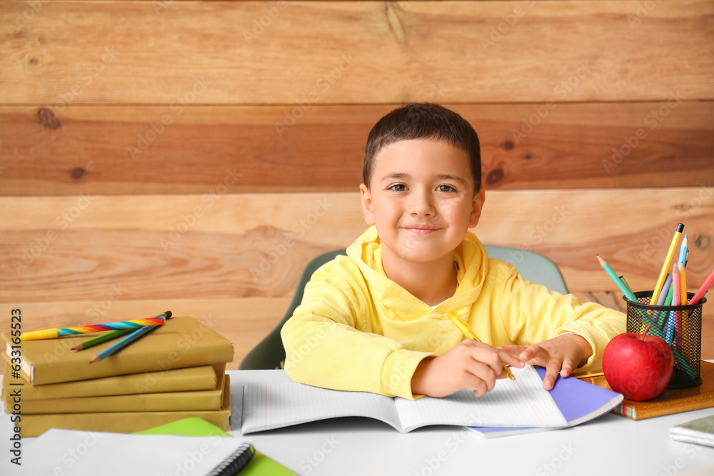 Cute little boy doing homework at table near wooden wall