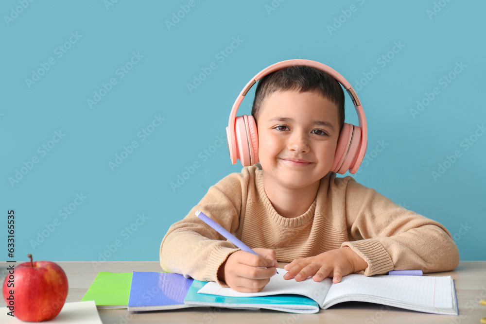 Cute little boy in headphones doing homework at table near blue wall