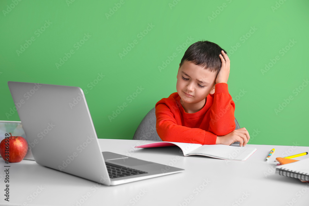 Stressed little boy doing homework at table near green wall