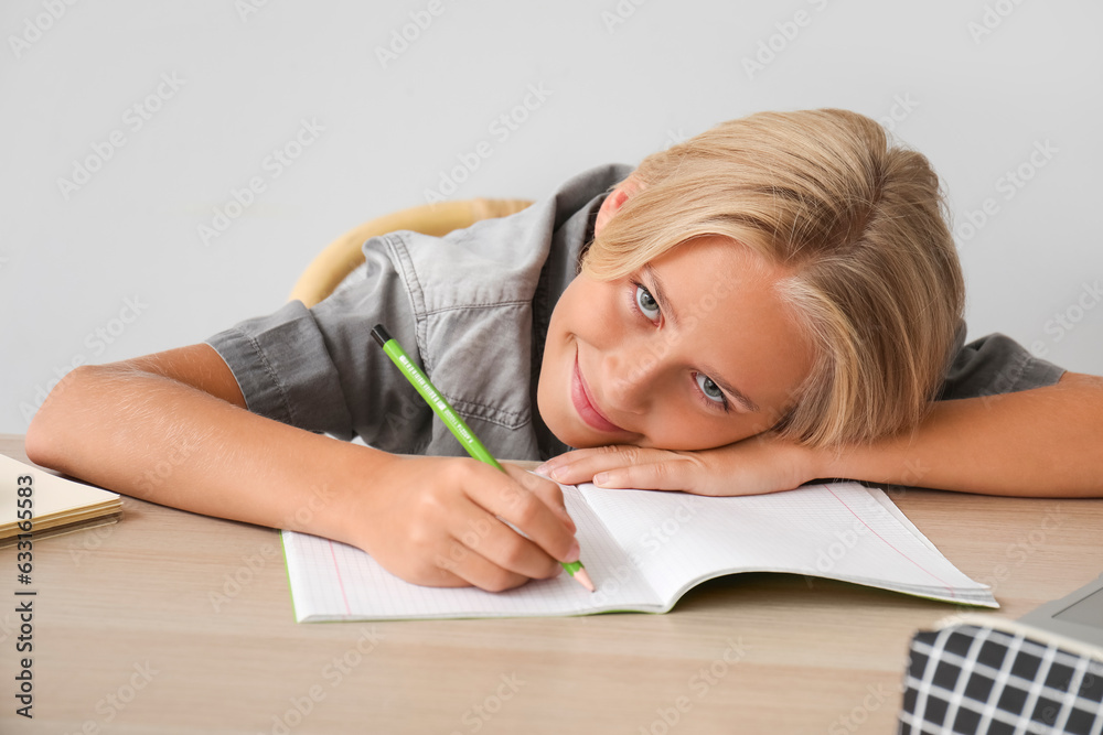 Little boy doing homework at table near light wall