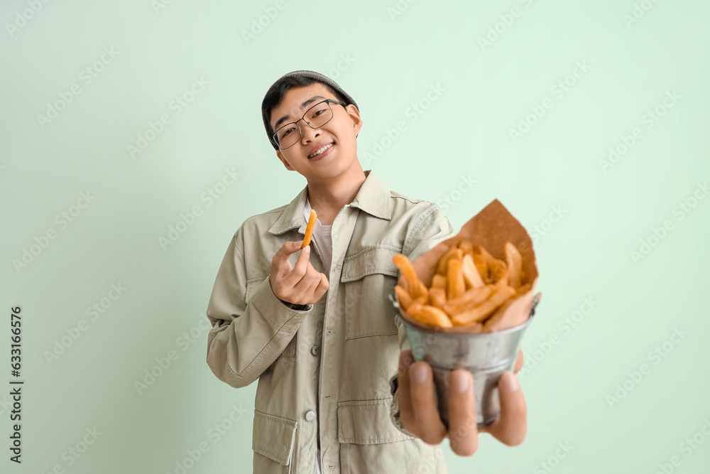 Young Asian man with bucket of french fries on green background