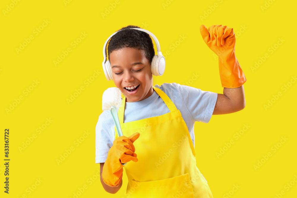 Cheerful African-American boy in headphones singing and using brush as microphone on yellow backgrou