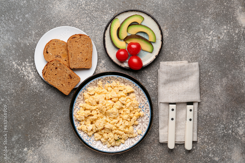 Plates of tasty scrambled eggs, bread, avocado and tomatoes on grey background