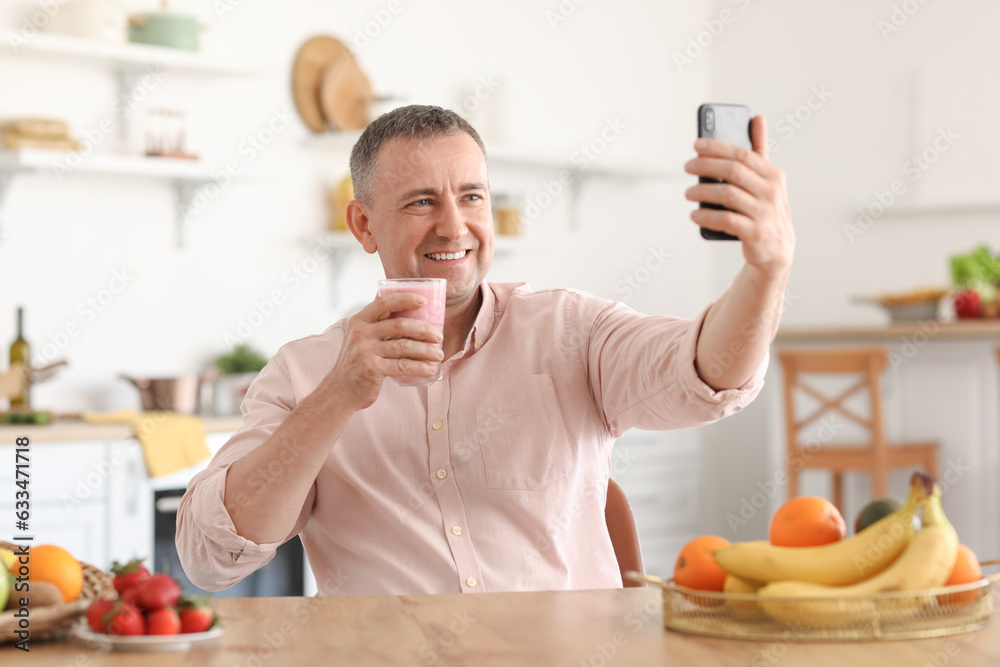 Mature man with glass of fresh fruit smoothie taking selfie in kitchen