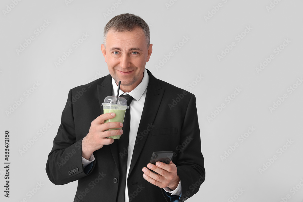 Mature businessman with mobile phone drinking vegetable smoothie on light background