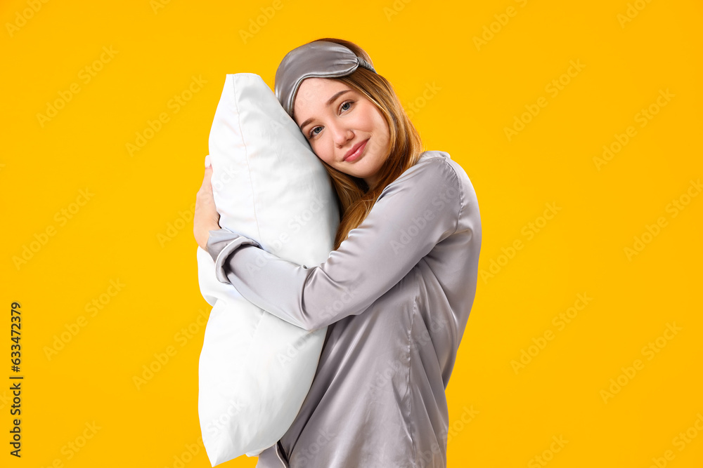 Young woman in pajamas with pillow on yellow background