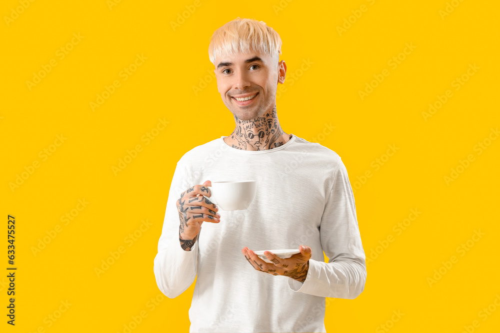 Young tattooed man with cup of coffee on yellow background