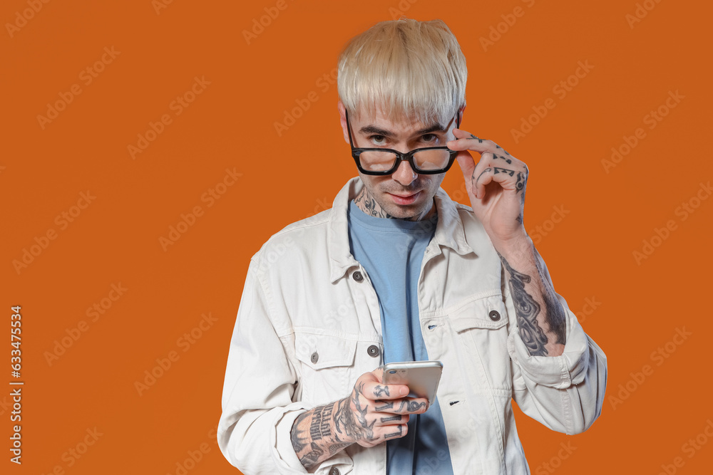 Young tattooed man in eyeglasses using mobile phone on color background