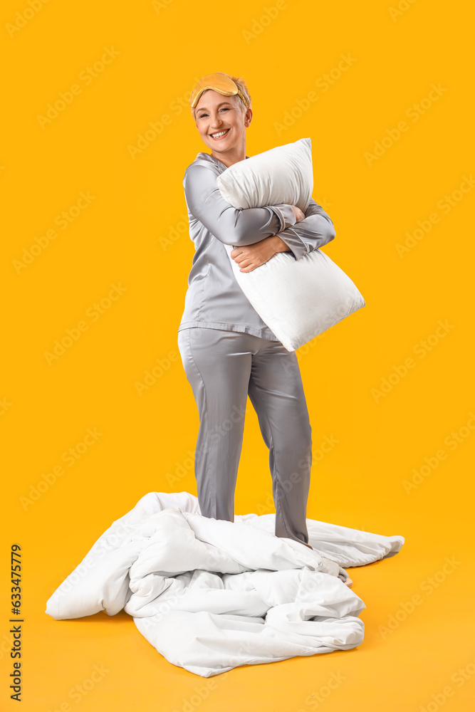 Mature woman in pajamas with pillow on yellow background