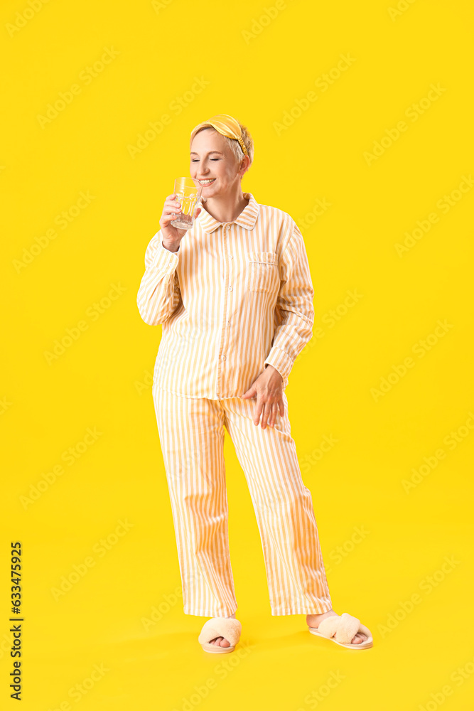 Mature woman in pajamas with glass of water on yellow background