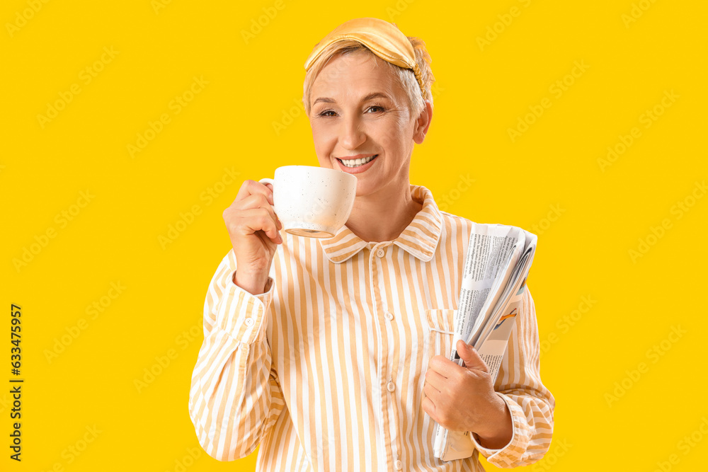 Mature woman in pajamas with cup of coffee and newspaper on yellow background