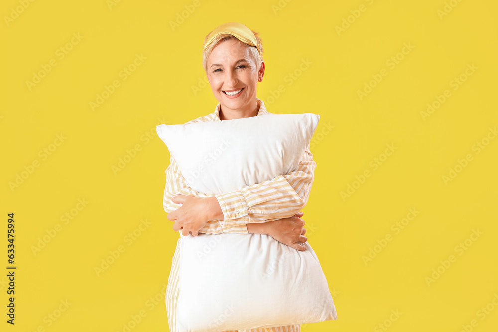 Mature woman in pajamas with pillow on yellow background