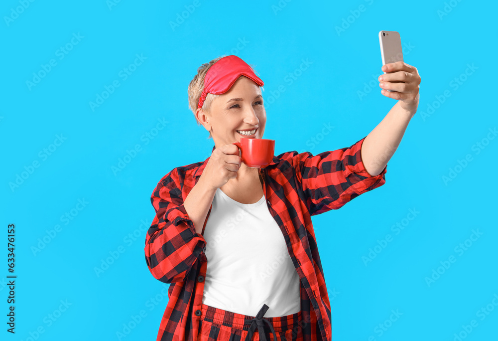 Mature woman in pajamas with cup of coffee taking selfie on blue background