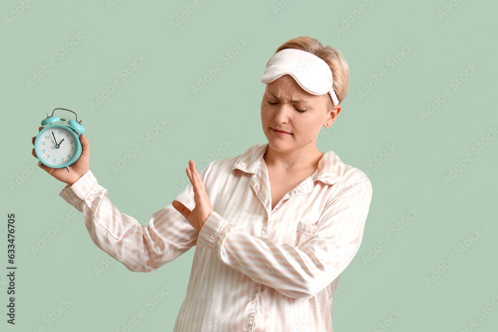 Displeased young woman in pajamas rejecting alarm clock on green background