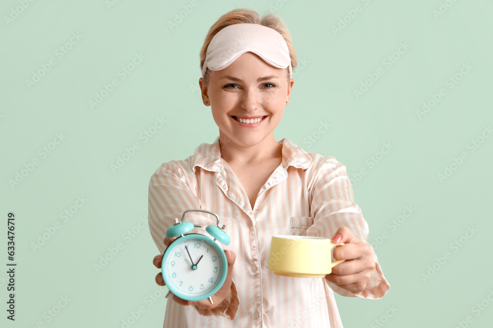 Young woman in pajamas with alarm clock and cup of coffee on green background