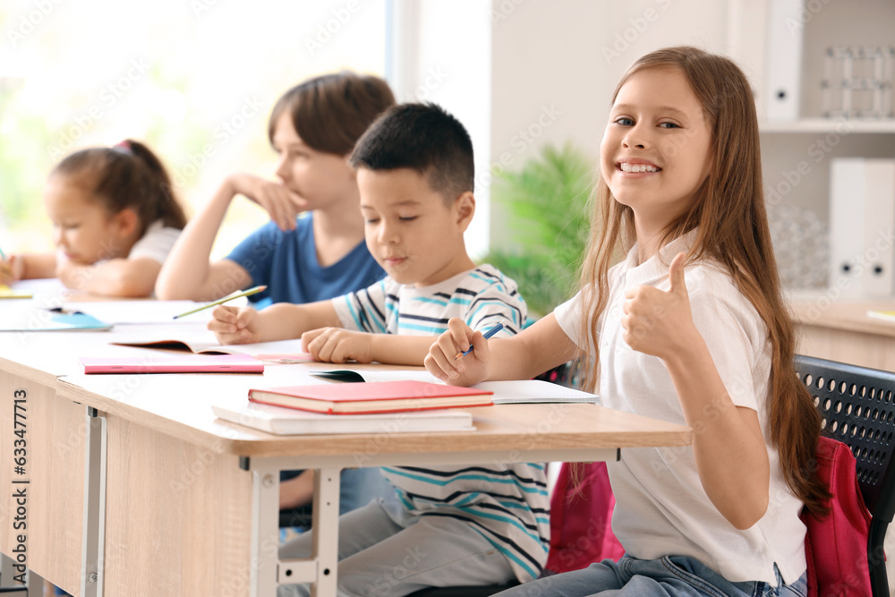 Little schoolgirl showing thumb-up during lesson in classroom