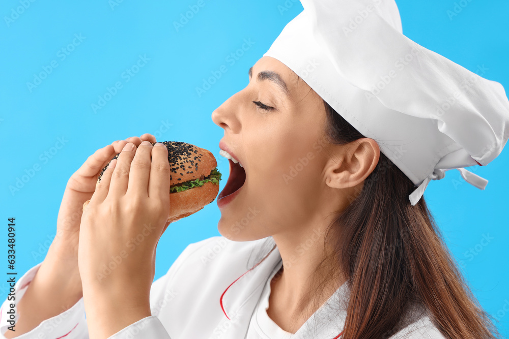 Female chef eating tasty burger on blue background, closeup