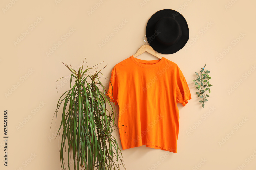 Stylish orange t-shirt and hat hanging on beige background