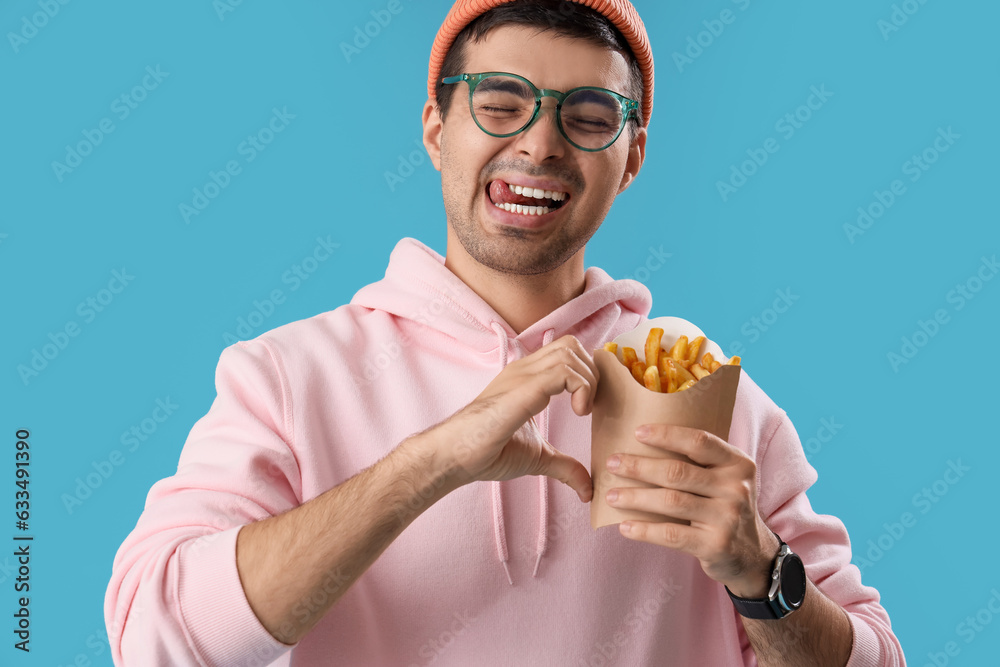 Young man with french fries making heart gesture on blue background, closeup