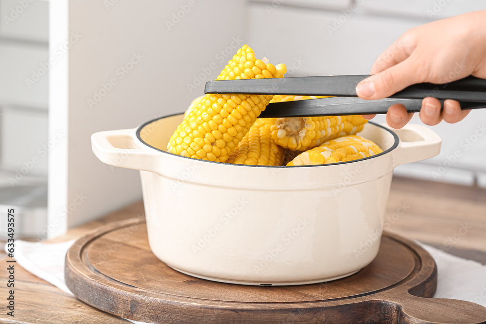 Woman taking out boiled corn cobs from cooking pot on wooden table in kitchen