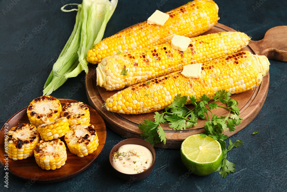 Wooden board and plate of tasty grilled corn cobs with butter on dark background