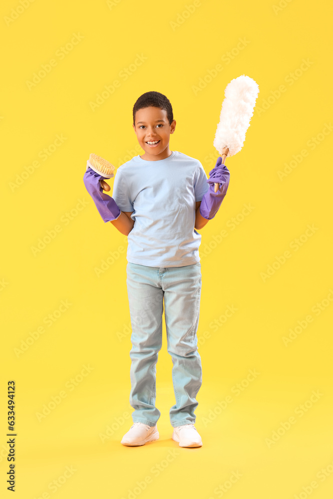 Cute little African-American boy with cleaning supplies on yellow background