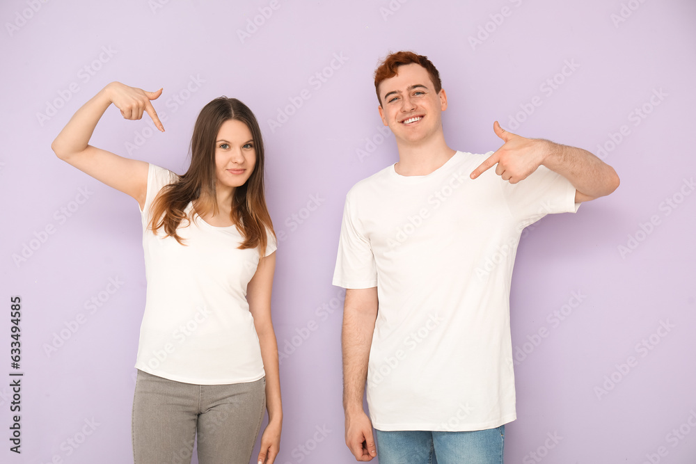 Young couple pointing at their white t-shirts on lilac background