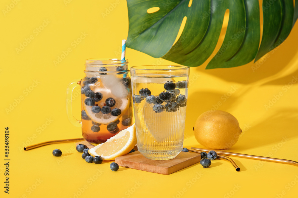 Glass and mason jar of fresh blueberry lemonade with monstera leaf on yellow background