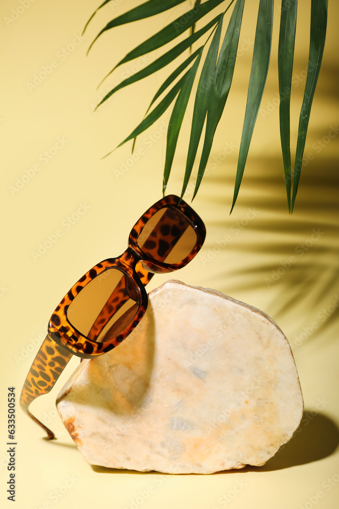 Stone with stylish sunglasses and palm leaf on yellow background