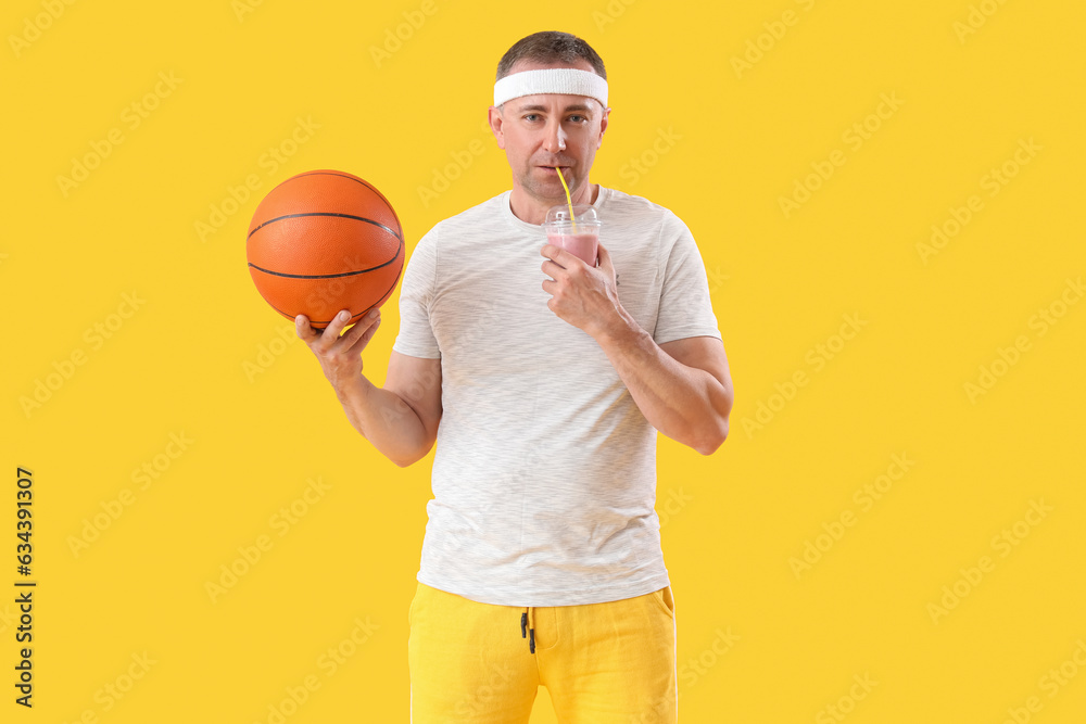 Sporty mature man with ball drinking smoothie on yellow background