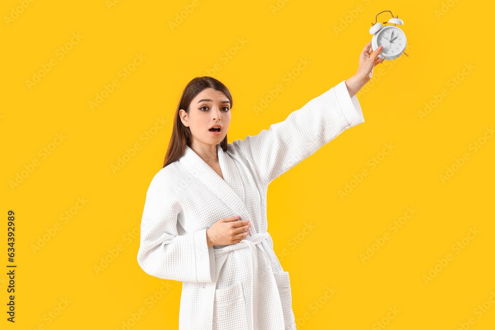 Shocked young woman with alarm clock on yellow background